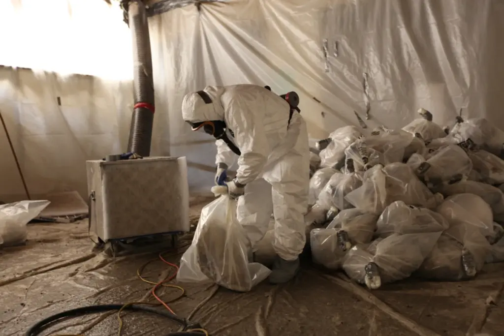 Operario con mono desechable y mascarilla embolsando residuos de amianto en una zona confinada con plástico
