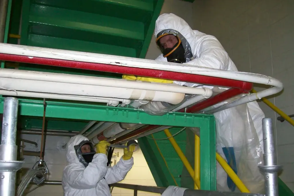 Operarios con mono desechable y máscara de protección respiratoria retirando el aislamiento de unas tuberías